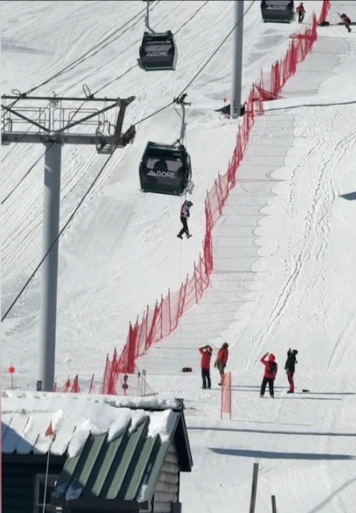 A person is being rescued from a ski gondola using a rope, with other people on the snowy ground below.