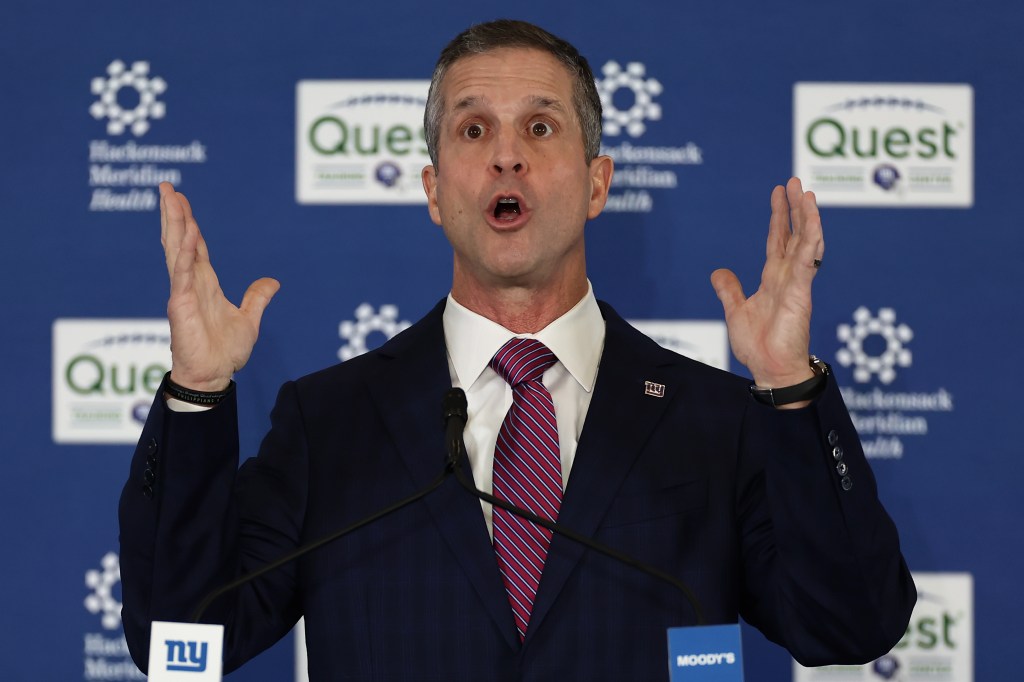 New York Giants head coach John Harbaugh speaking at a news conference, with his arms raised and mouth open.
