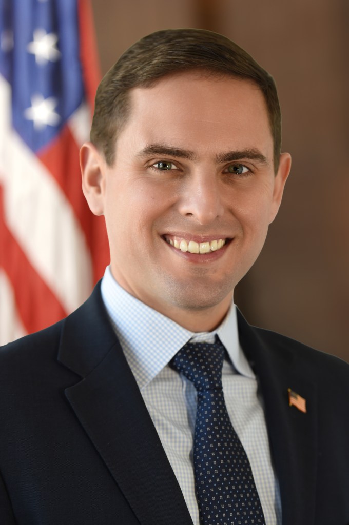 New York State Assembly Member Matt Slater smiling, wearing a dark suit, light blue patterned shirt, and dark blue tie with small light dots, with an American flag to his left.