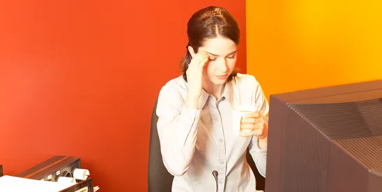 person sitting at desk in front of desktop computer, rubbing temples; NYC DCWP Publishes ESSTA Amendments FAQs and Form Notices