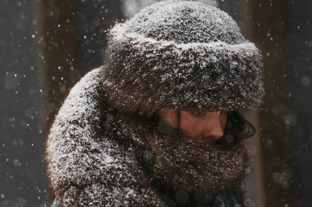 Pedestrian with long hair covered in snow, wearing a fur hat, during a winter storm.