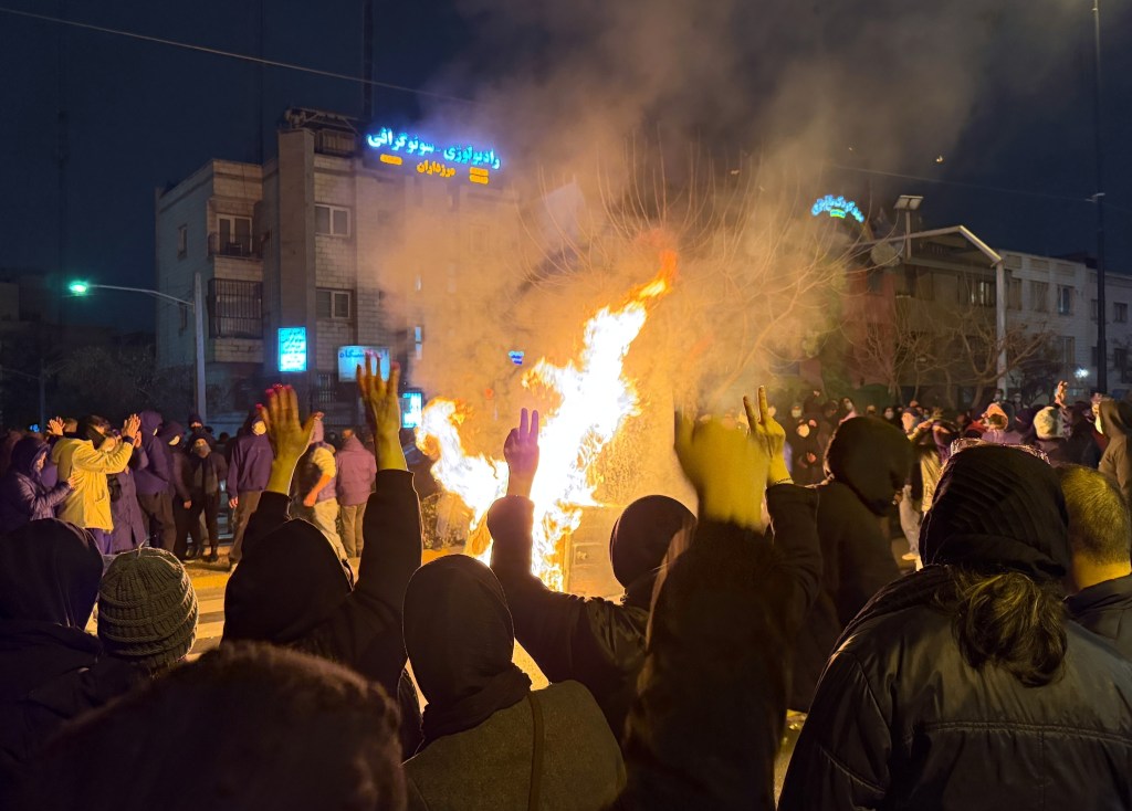 People raising their hands in protest around a large bonfire at night in Tehran.