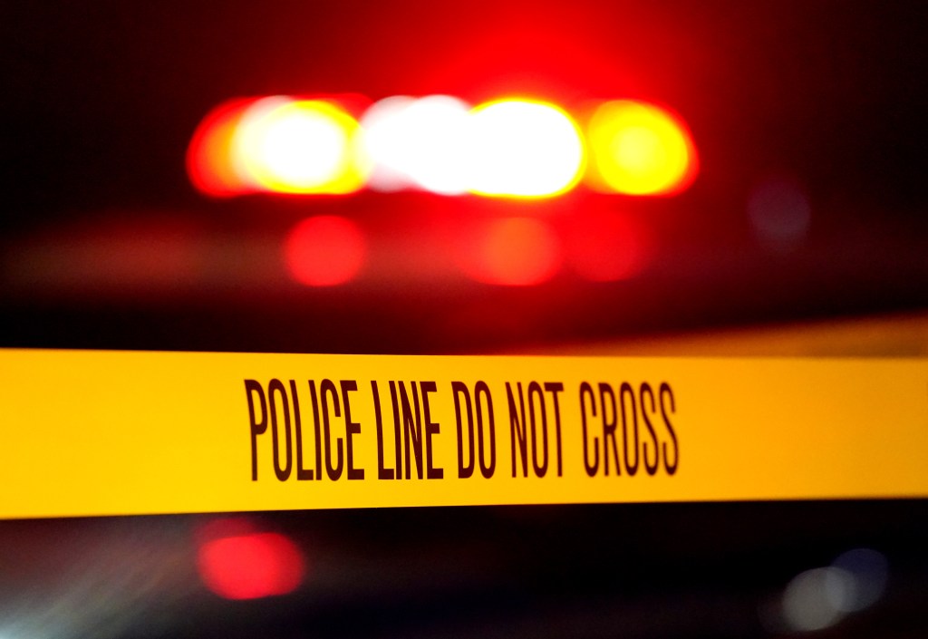 Police Line Do Not Cross tape at a crime scene with flashing red lights in the background.