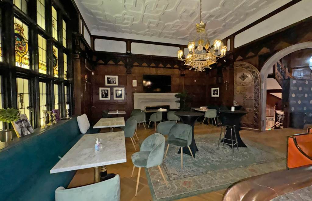 The interior of the Friars Club, a New York establishment, featuring dark wood paneling, stained glass windows, a chandelier, and a mix of tables and chairs.