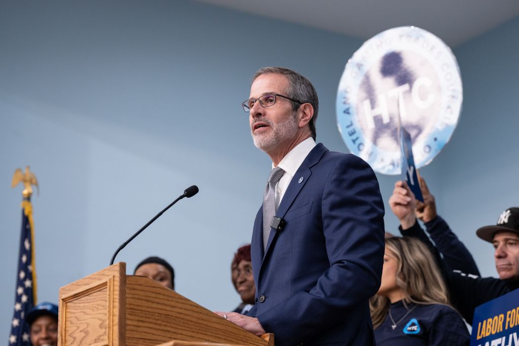 Rich Maroko, President of the Hotel and Gaming Trades Council (HTC), speaks at a podium with an HTC union sign and supporters behind him.