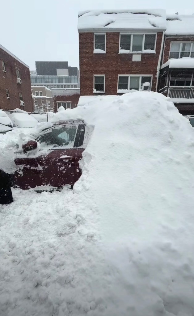 A red car buried under a large mound of snow on a city street.