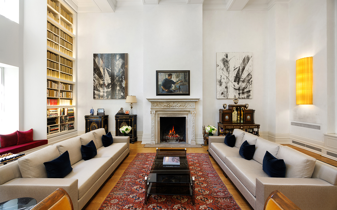 A spacious, elegant living room with two beige sofas facing each other, a red patterned rug, a wood-burning fireplace, bookshelves, modern art on the walls, and large windows letting in natural light.