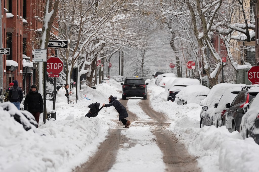 A woman pushes a stroller through plowed snow on a street lined with snow-covered cars and trees.