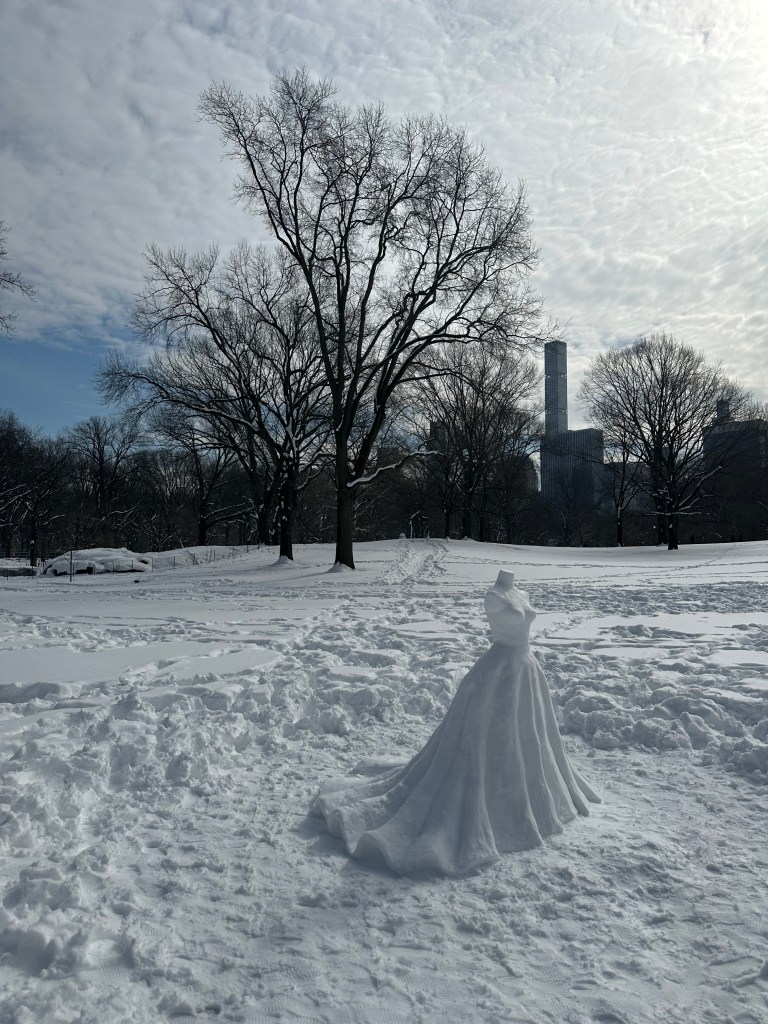 A snow mannequin dressed in a realistic snow gown stands in a snow-covered park with trees and city buildings in the background.