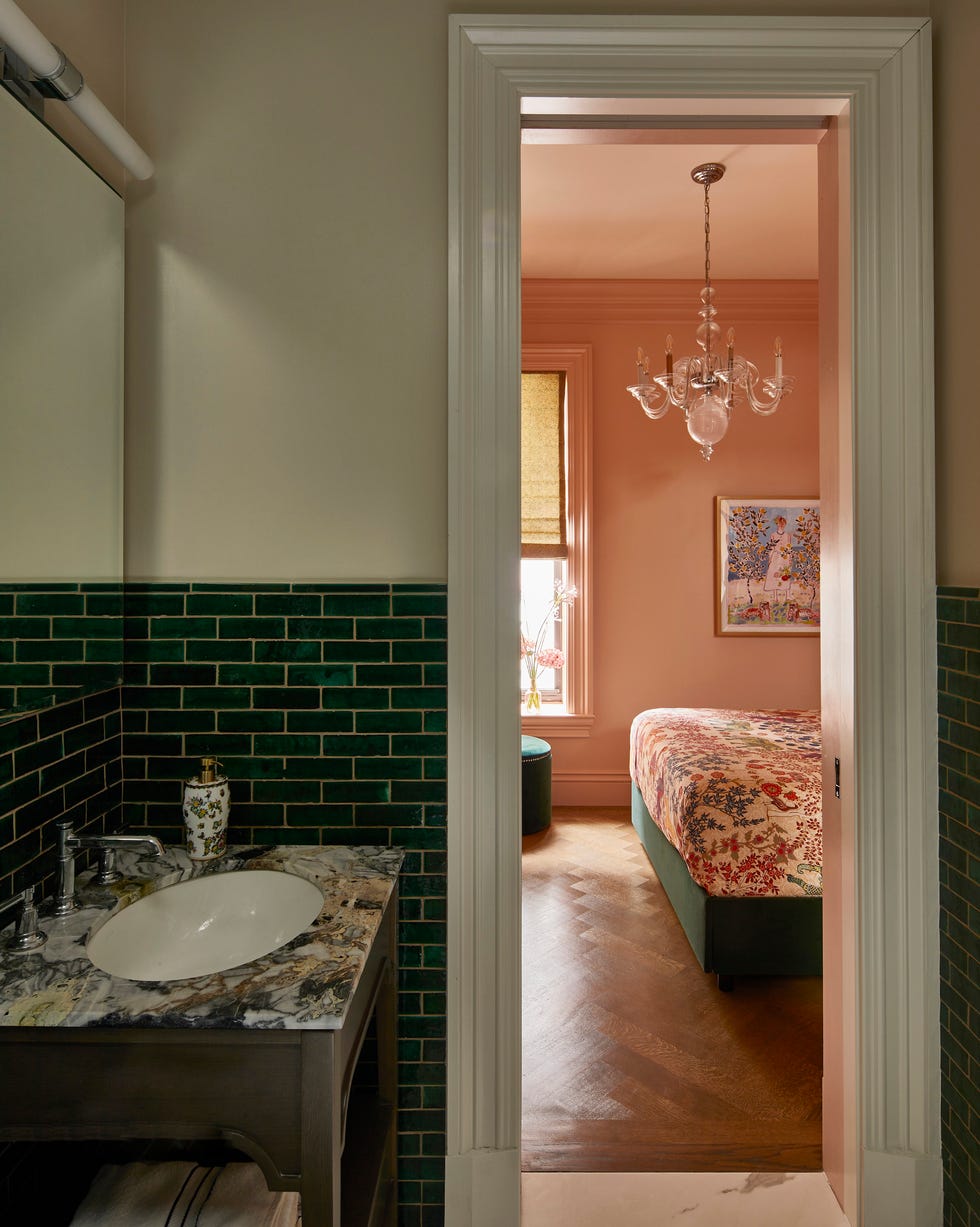 view from a bathroom with green tile walls and a vwood vanity with stone countertop into a bedroom with a chandelier