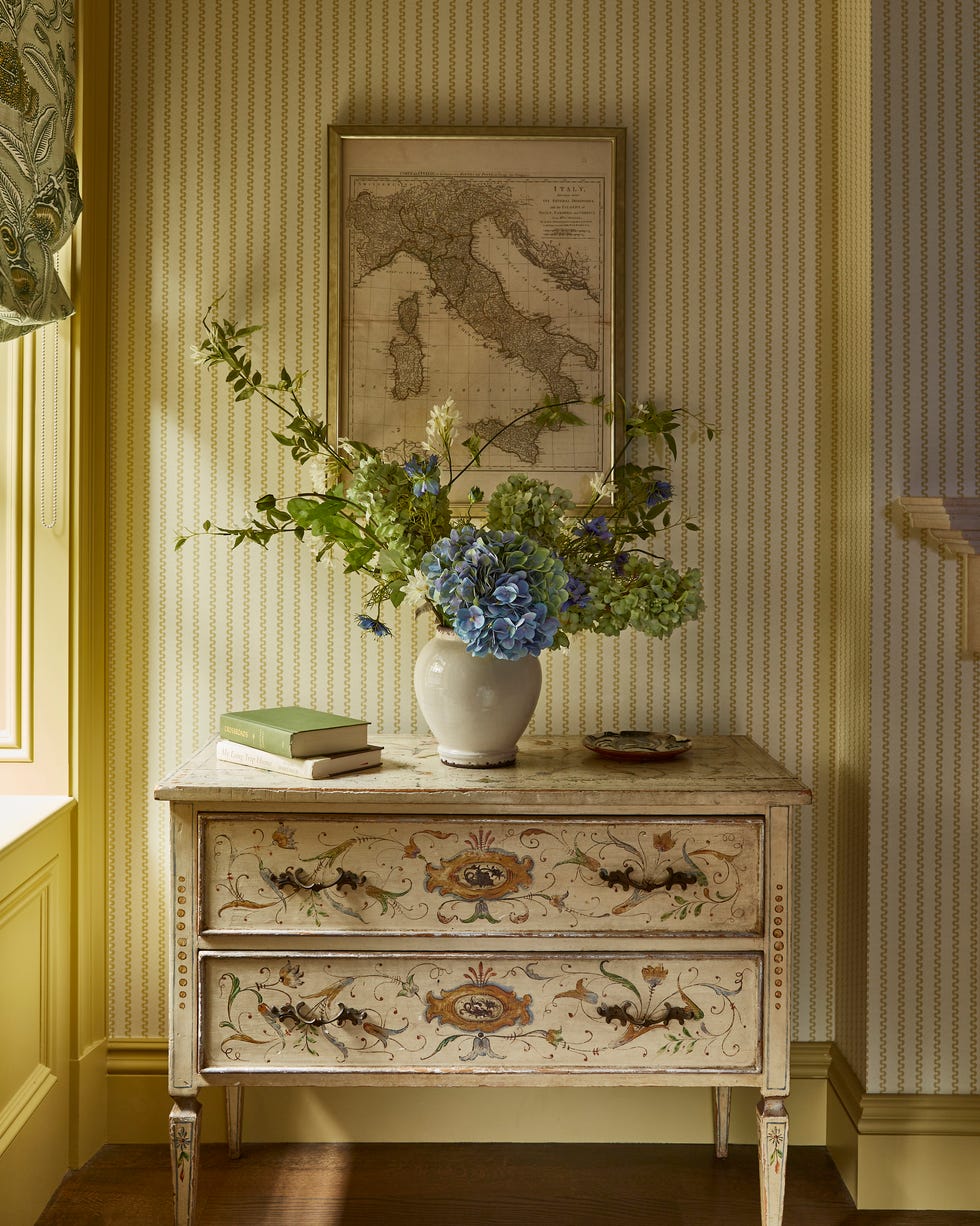 chest with decorative paint with a floral arrangement on top and a framed map above it in front of yellow striped wallpaper