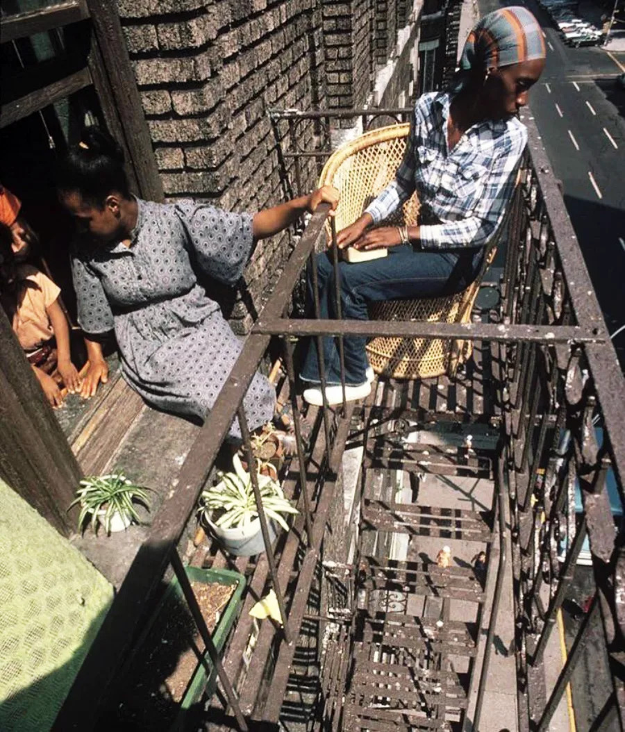 Two women sit on a fire escape outside a brick building; one is in a wicker chair, the other by an open window. Potted plants are nearby, and a city street with cars is visible below.