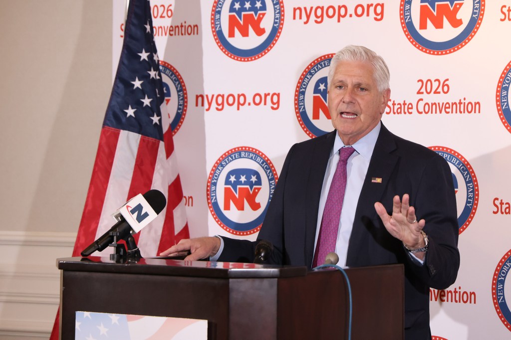 Bruce Blakeman speaking at a podium with an American flag and New York Republican Party banners in the background.