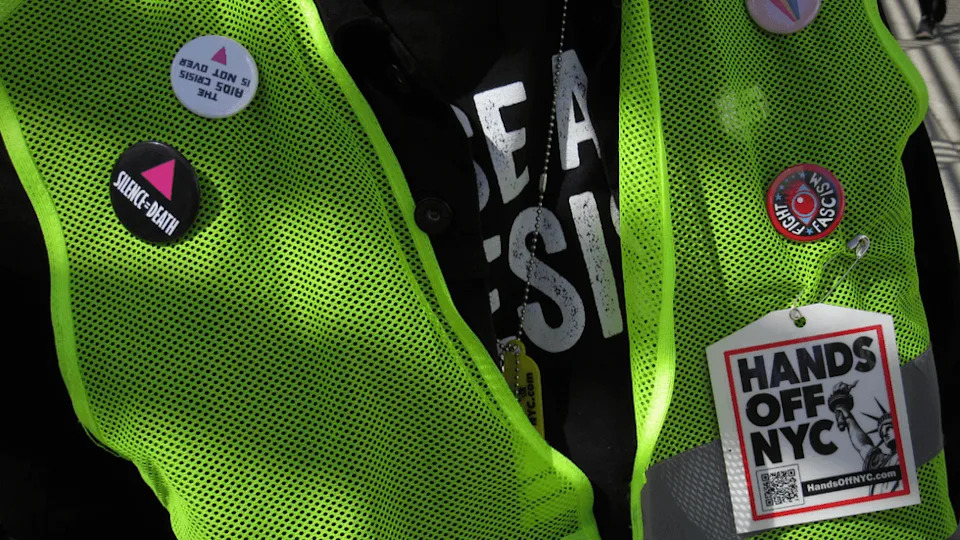 On a yellow, mesh vest, several activist pins and buttons are displayed. One reads "Silence = Death" with a pink triangle. Another reads "Hands Off NYC."