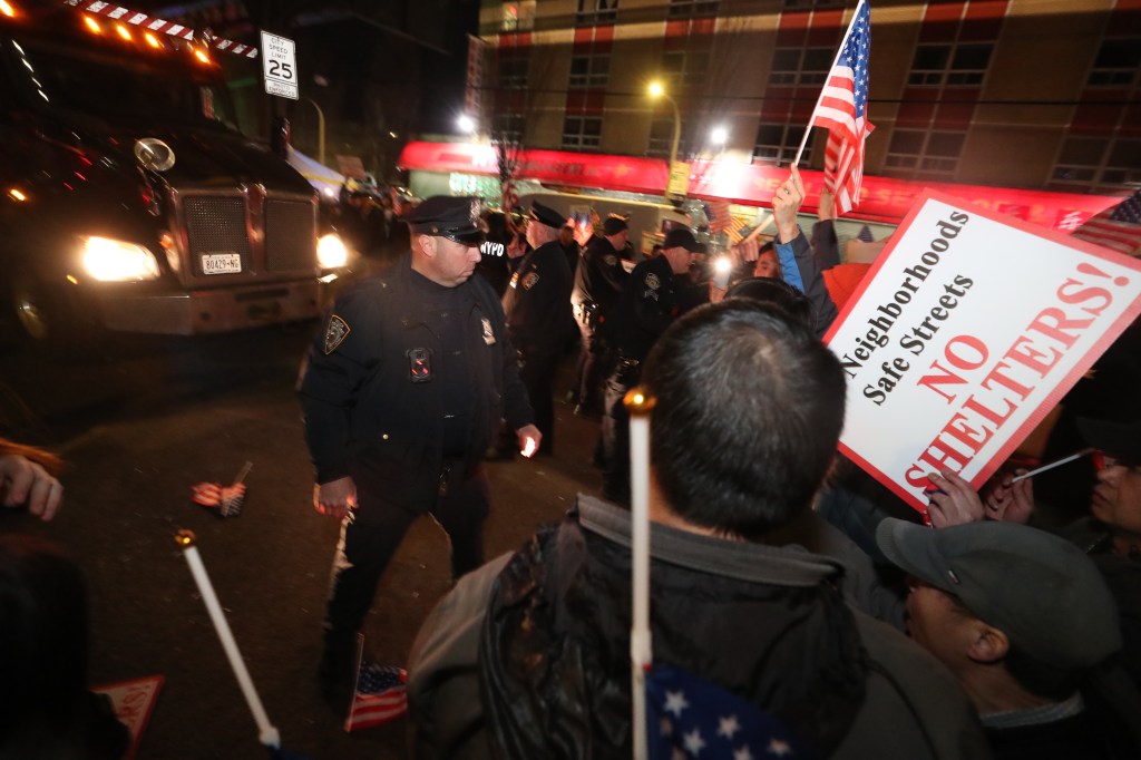 Police officers and protesters with an "NO SHELTERS!" sign gather around a construction vehicle at night.