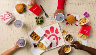 Overhead view of Chick-fil-A items, including chicken sandwiches, fries, chicken nuggets, and drinks, on a wooden table with visible hands interacting with the food.
