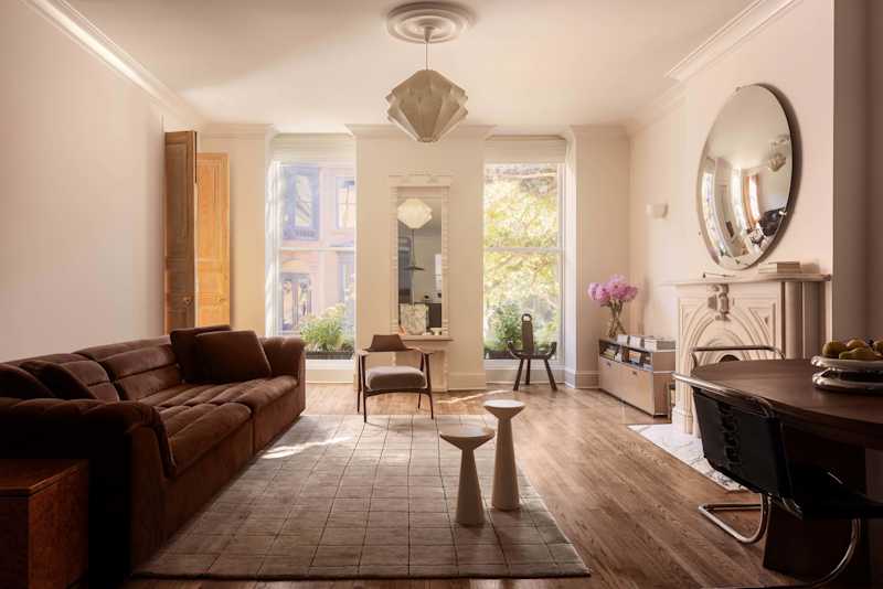Living room with a brown sectional sofa, two white side tables, a round mirror, and large windows with plants.