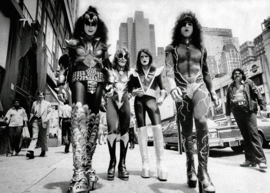 Four people in elaborate rock costumes and face paint walk down a busy city street, flanked by tall buildings and passing pedestrians; two wear platform boots, and one has lightning patterns on their outfit.