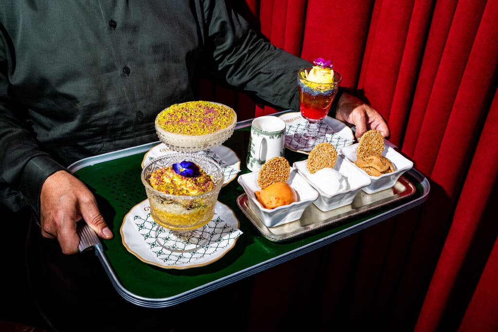 A man in a silky black shirt holds a tray of desserts. 