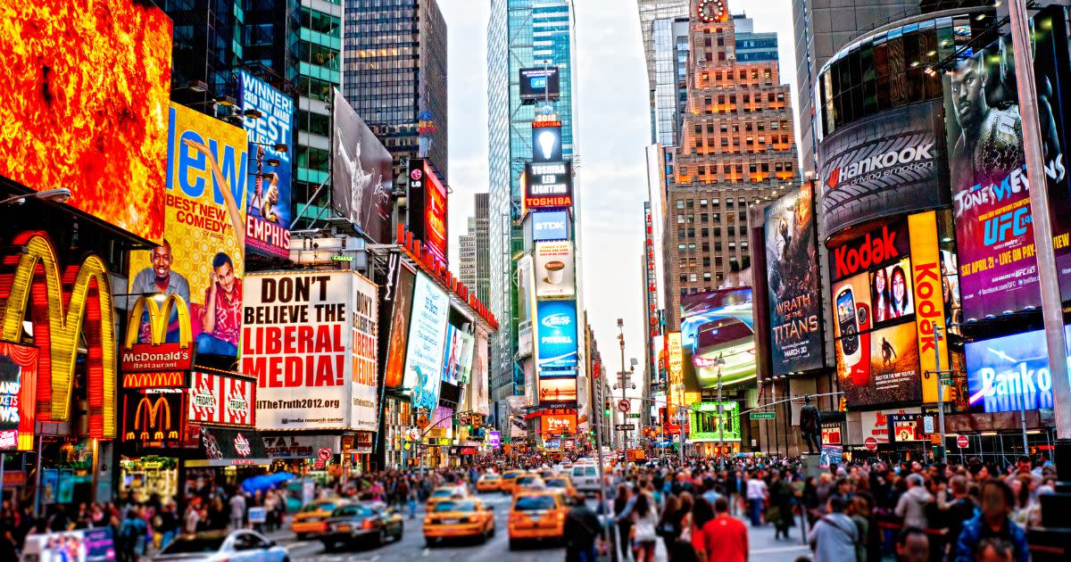 There's a vibrant panoramic, wide shot of New York City's "Times Square", and a multitude of people are walking on the streets as yellow cabs and cars move along the busy city streets.