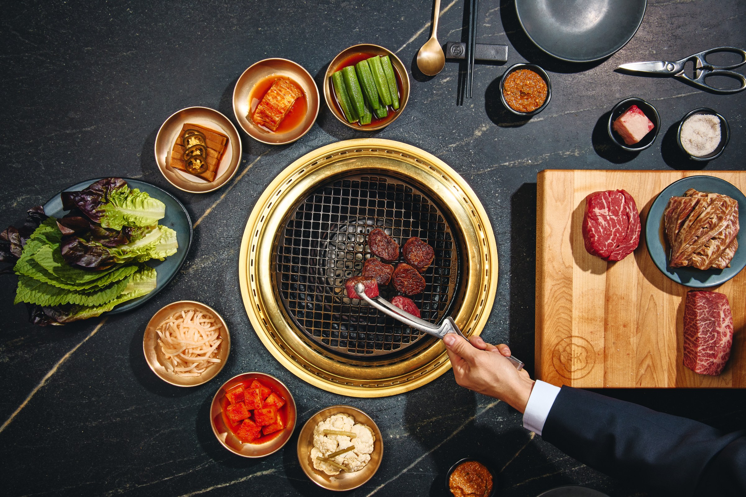 A table with someone grilling meat in the middle surrounded by plates of vegetables and a board of meat.
