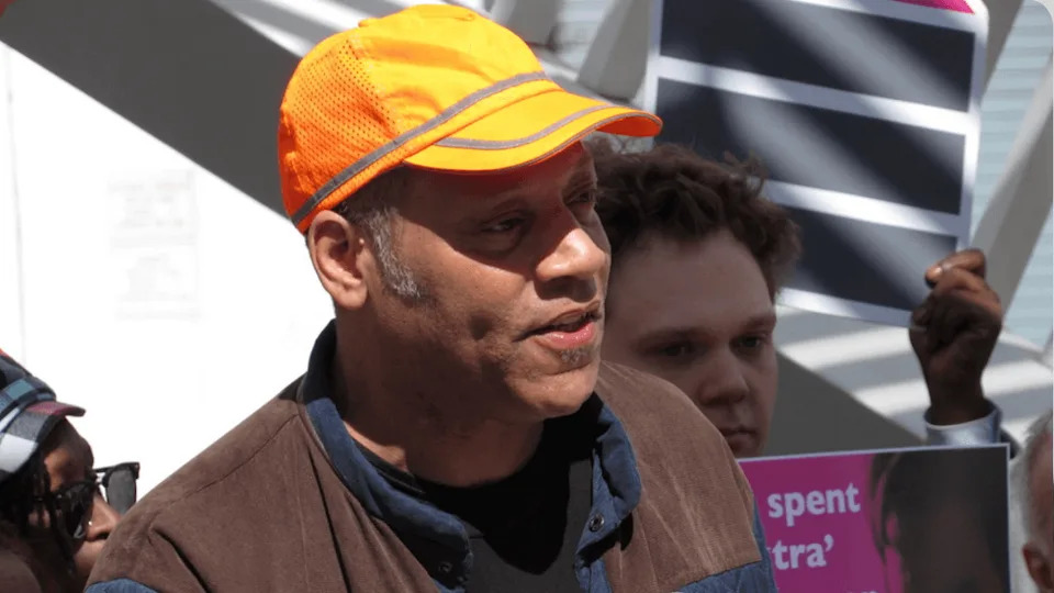 A person in a bright, orange hat speaks to a crowd.