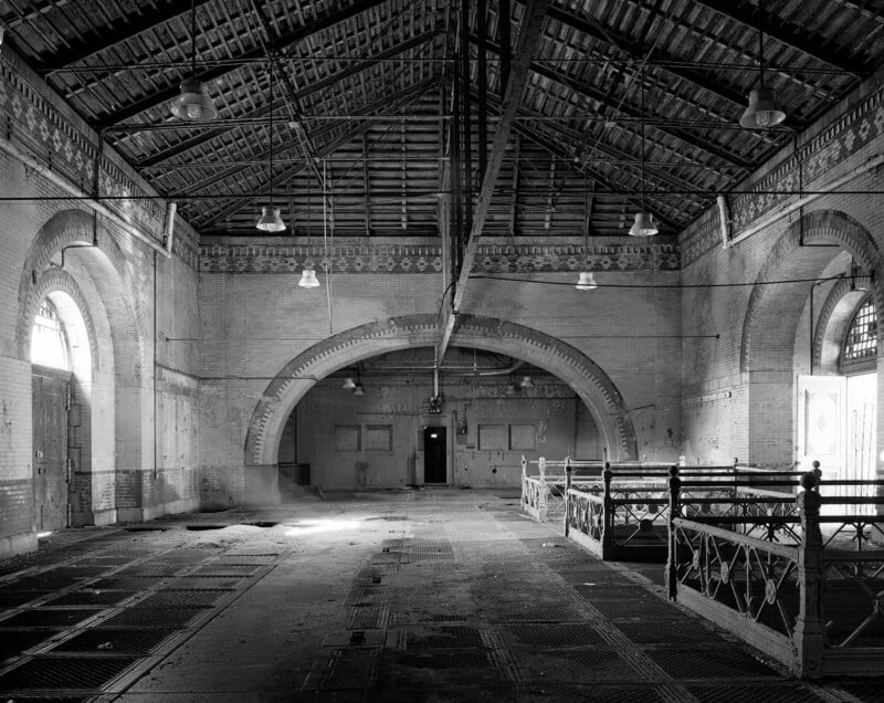 Black and white photo of a large, empty hall with high ceilings, arched windows, and a central doorway. The space has exposed beams, metal railings, and visible wear, giving an abandoned, industrial feel.