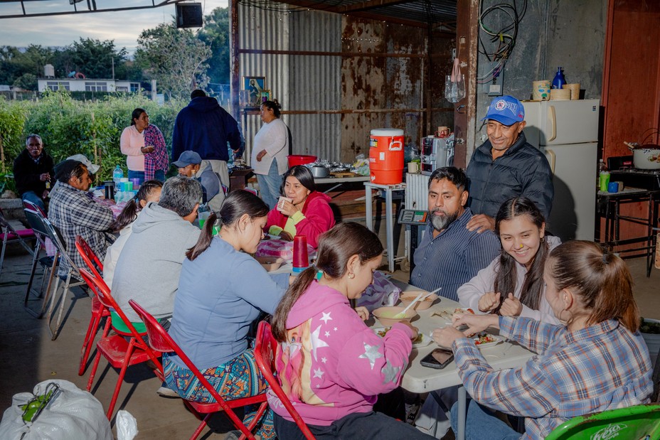 photo of family members sitting in folding chairs at a meal at long table inside corrugated metal structure