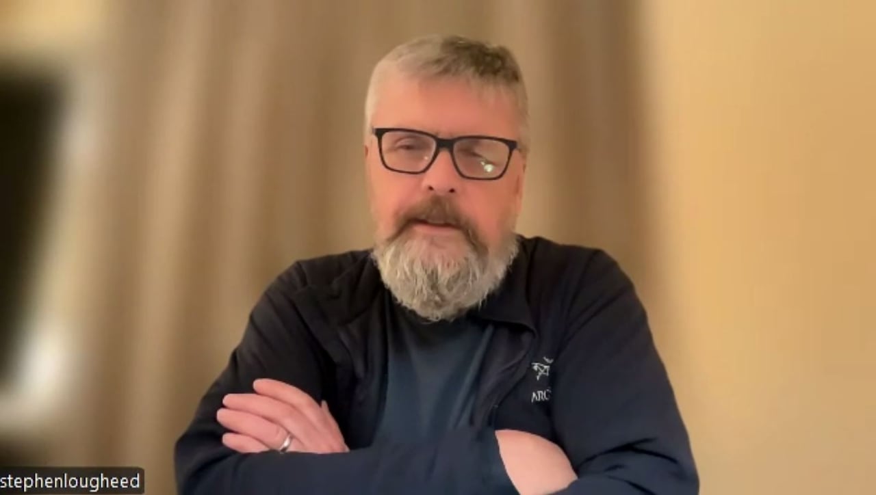 A man with glasses and a grey beard sits with crossed arms in a hotel room.