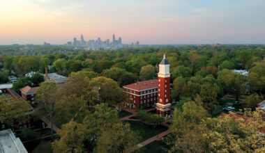 An aerial view of Queens University’s campus shows the clock tower rising above trees, with the Charlotte skyline in the distance.