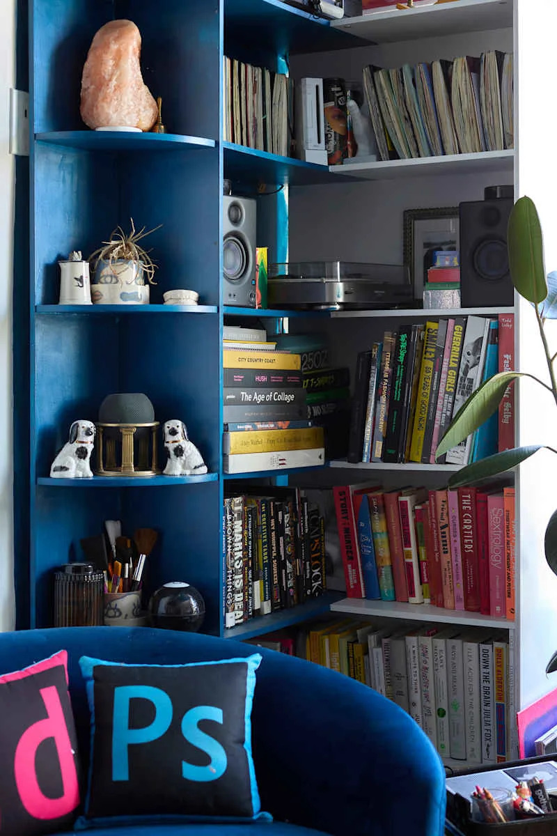 Bookshelves filled with colorful books, a record player, decorative items, and a blue velvet sofa with vibrant pillows.