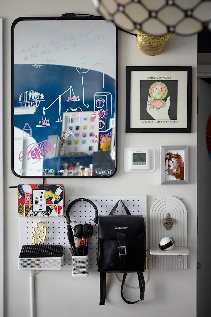 A wall display featuring a mirror with colorful doodles, framed art, a thermostat, and organized accessories like a backpack, headphones, and pens.