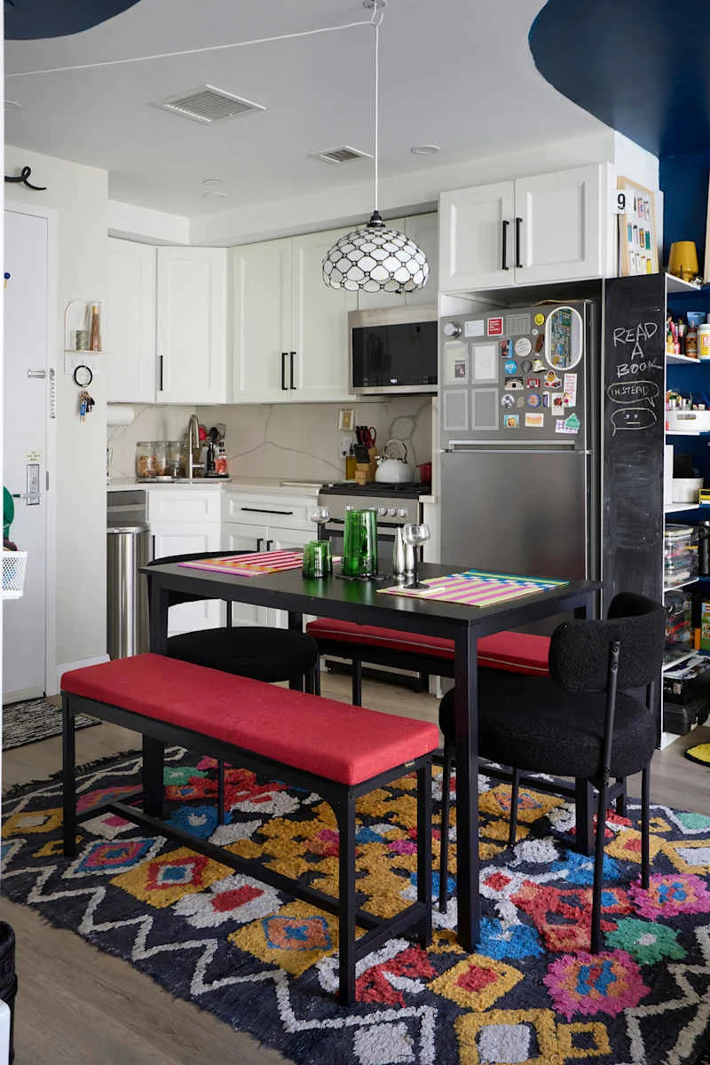 Modern kitchen with white cabinets, a black and white pendant light, red benches, and a colorful patterned rug.