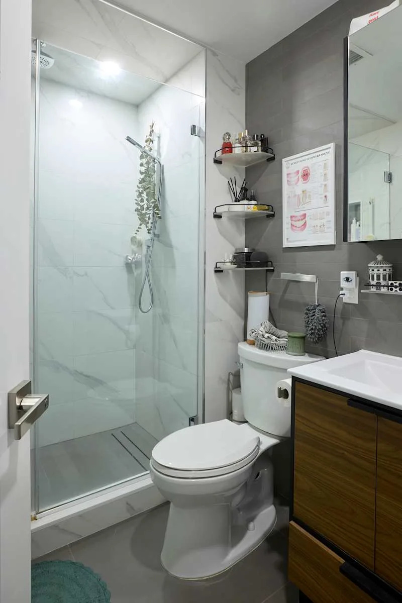 Modern bathroom featuring a glass shower, toilet, and wooden vanity with storage. Shelves hold toiletries and decor.