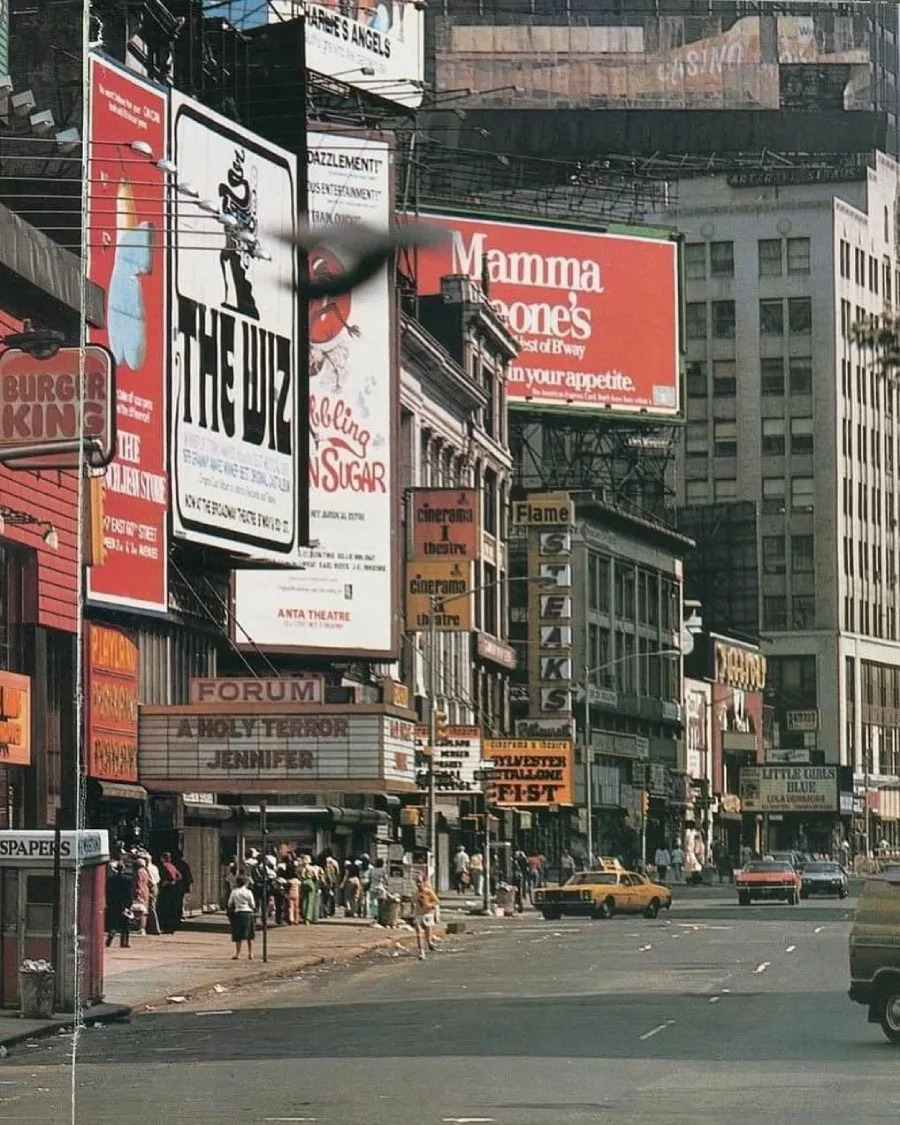 A busy city street lined with theaters, restaurants, and billboards, including ads for Burger King, Mamma Leone’s, The Wiz, and Sugar Babies. People walk on the sidewalks and cars drive by in daylight.