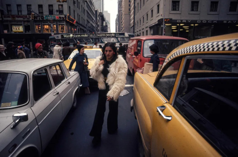 A person in a white fur coat walks between closely packed cars, including a yellow taxi, on a busy city street filled with people and traffic. Tall buildings line both sides of the street.