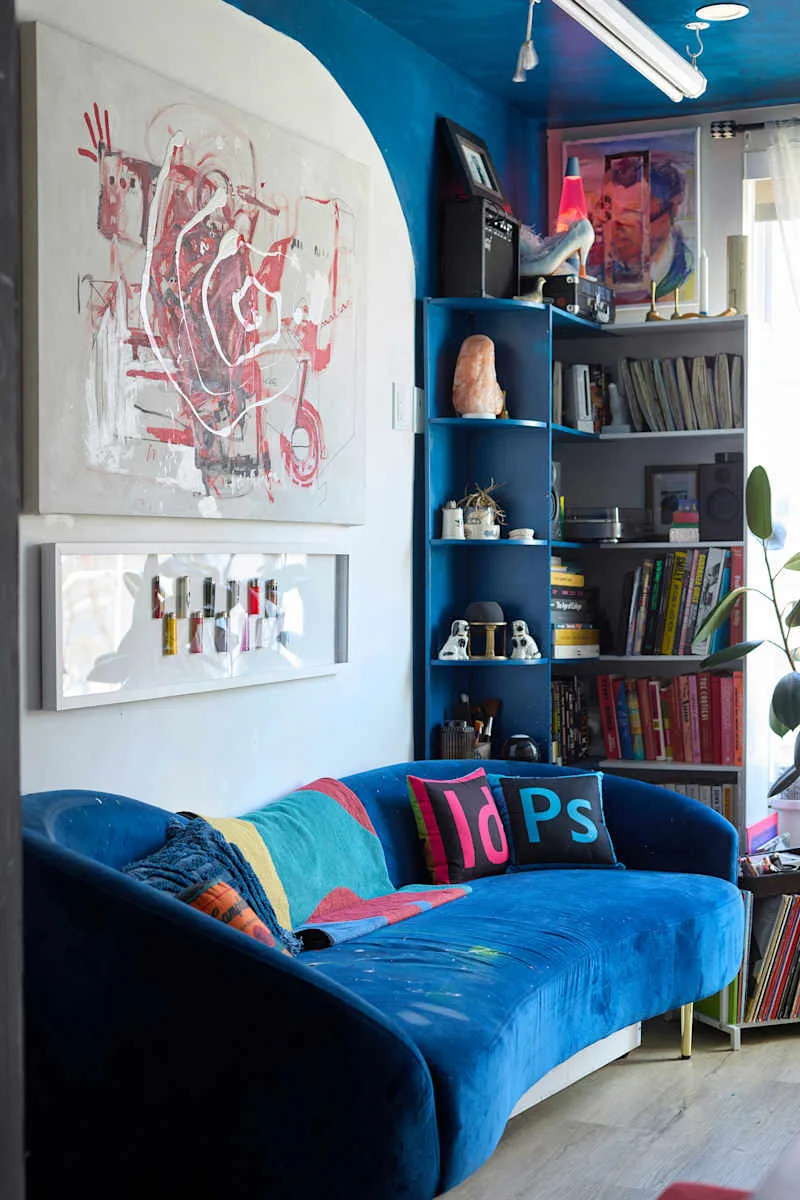 A vibrant blue curved sofa with colorful pillows, surrounded by bookshelves, artwork, and decor items.