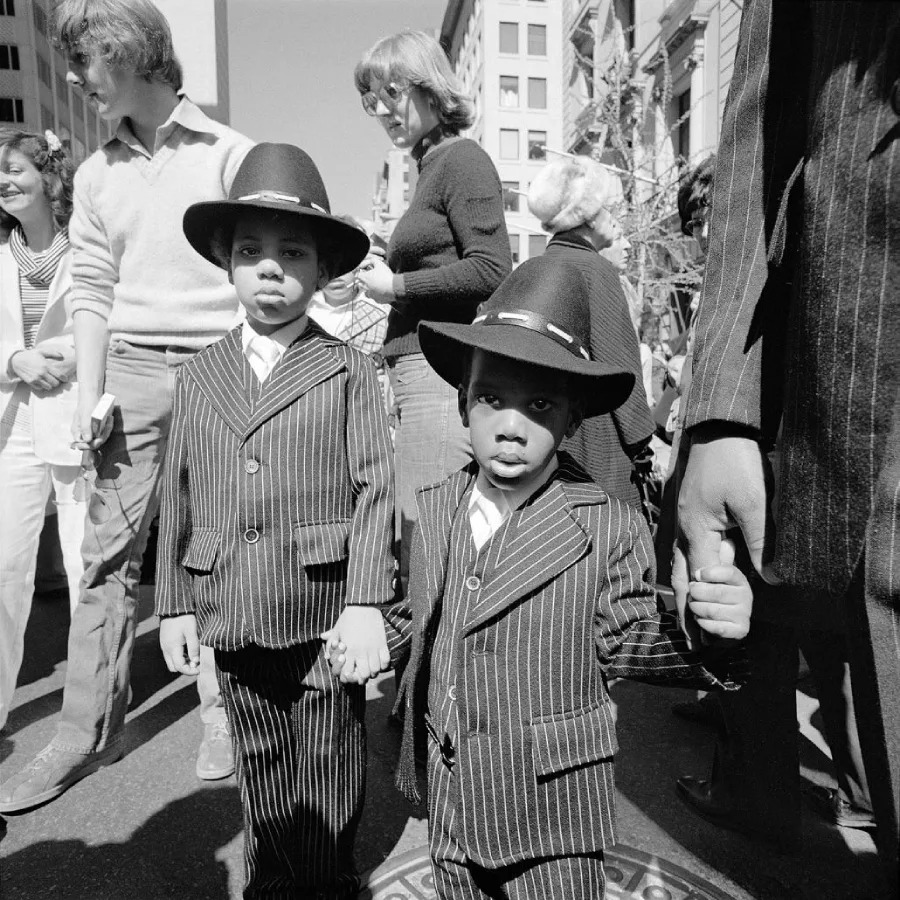 Two young children wearing pinstripe suits and wide-brimmed hats hold hands while standing in a crowd on a city street during daylight. Adults and other people walk and stand around them in the background.