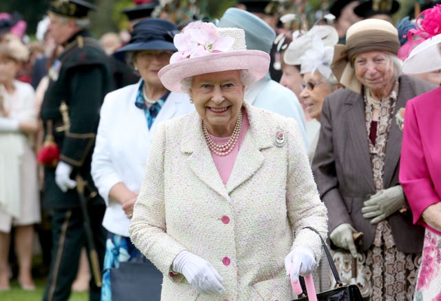 The late Queen smiling while wearing a white coat and pink dress, with matching pink hat, during a garden party