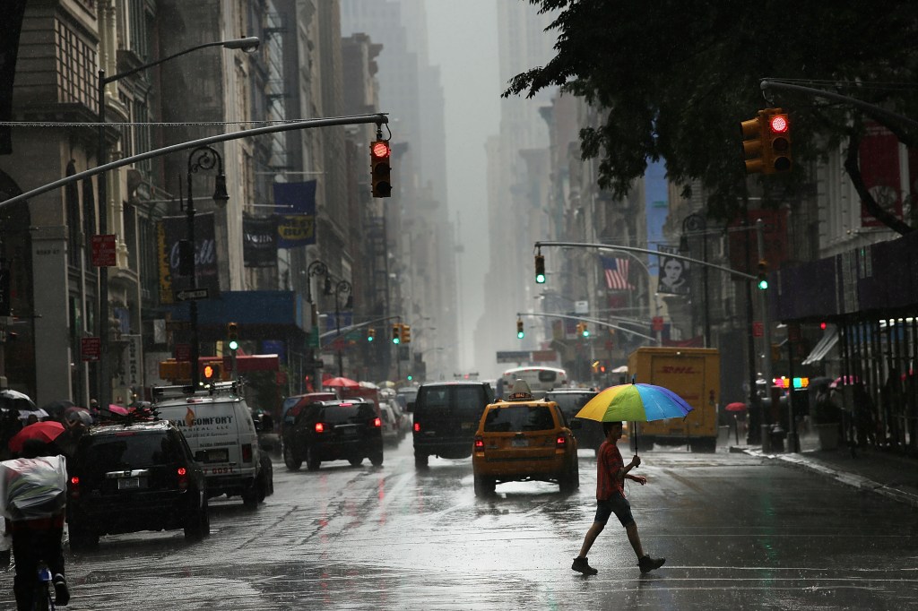 A man carrying a rainbow-colored umbrella walks across a rainy New York City street with taxis and other cars in the background.
