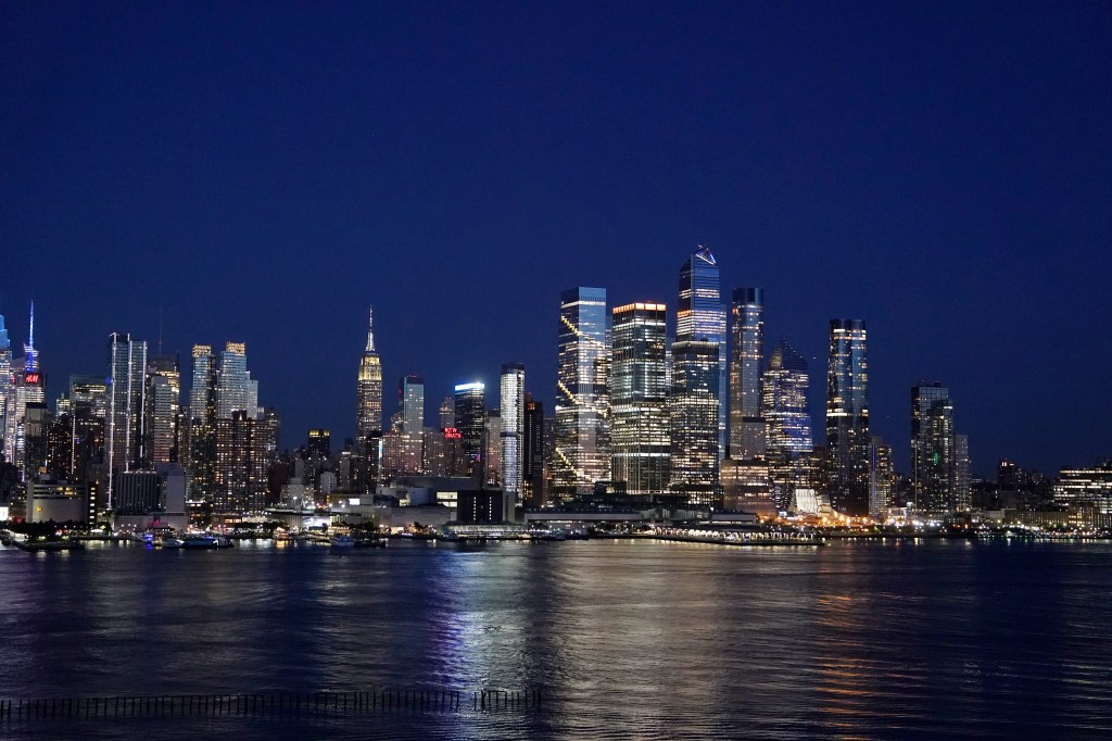 New York City skyline at night from Weehawken, NJ, with reflections of buildings on the water.