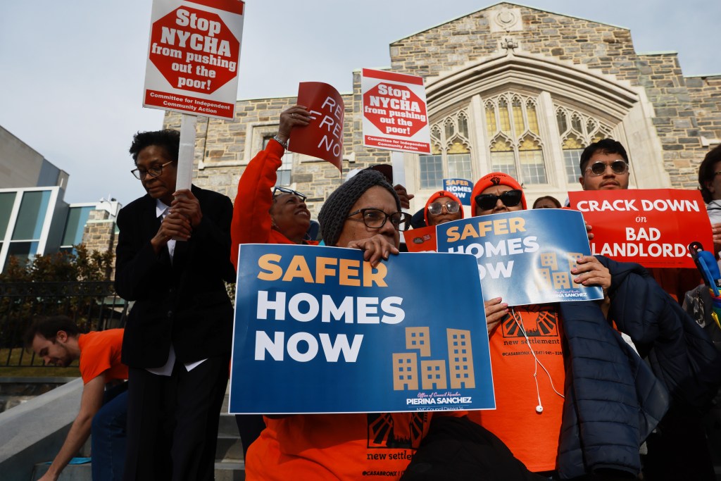 People hold signs that read "Safer Homes Now" and "Stop NYCHA from pushing out the poor!" at a rental ripoff hearing.