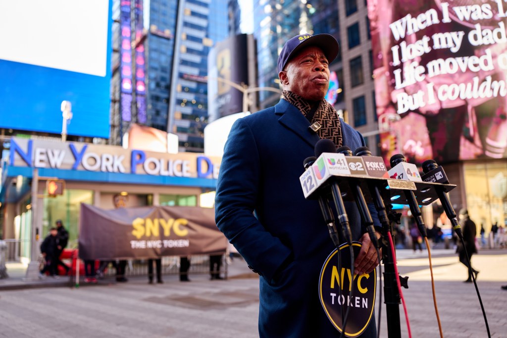 Former NYC Mayor Eric Adams speaking at a press conference in Times Square, holding an "NYC TOKEN" sign.