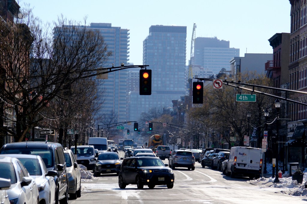 Traffic flows along Washington Street in Hoboken, New Jersey, with tall buildings in the background and red traffic lights overhead.
