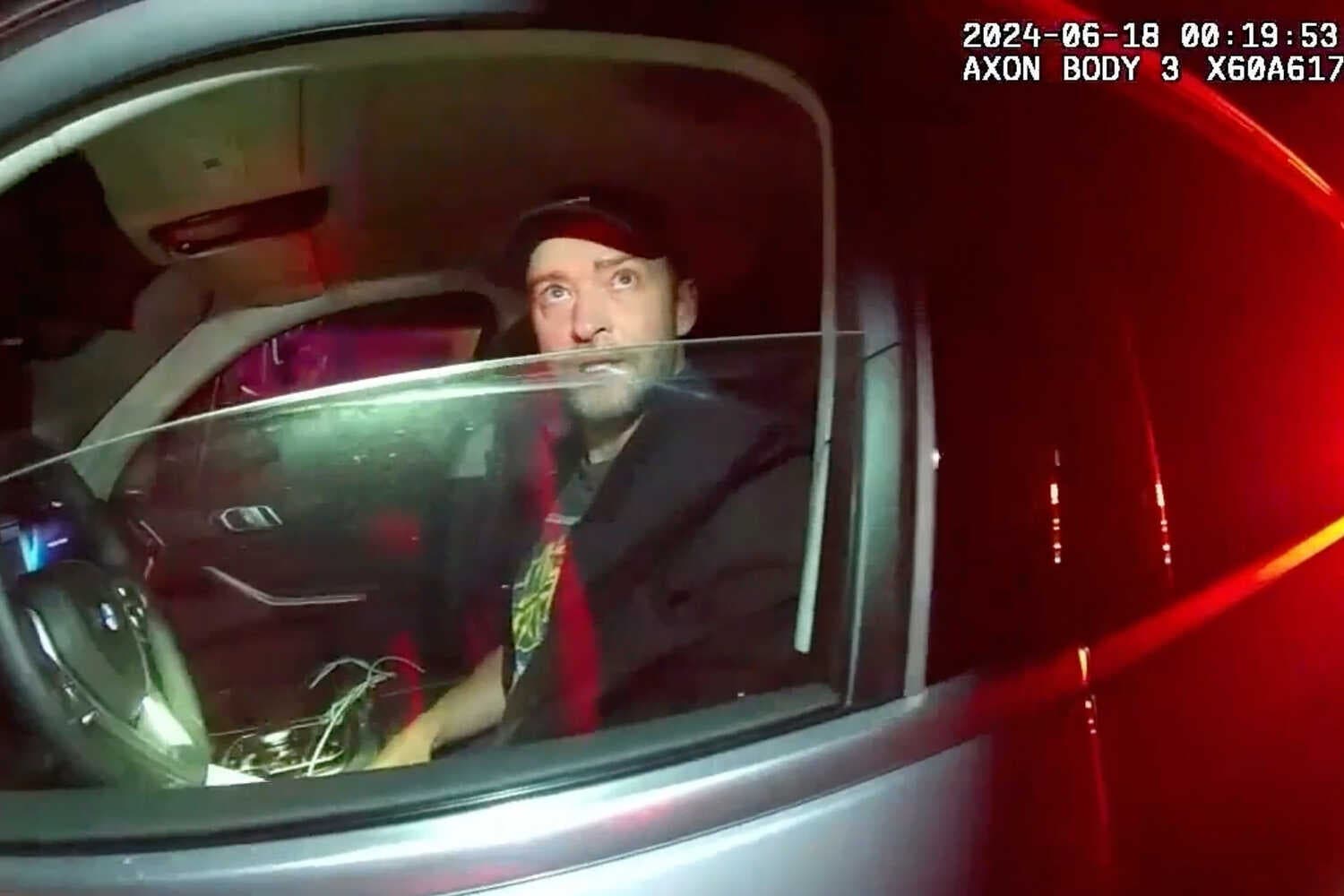 Justin Timberlake, wearing a baseball cap, speaks to a police officer from the driver’s seat of his car. 