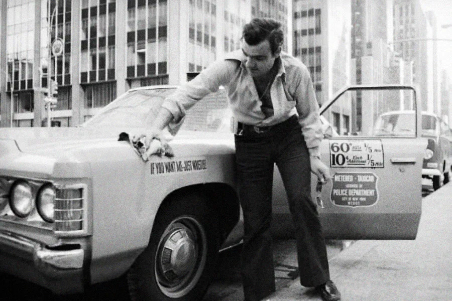A man in a collared shirt and slacks cleans the side of a taxi parked on a city street, with tall office buildings in the background and a taxi fare sign on the car door.