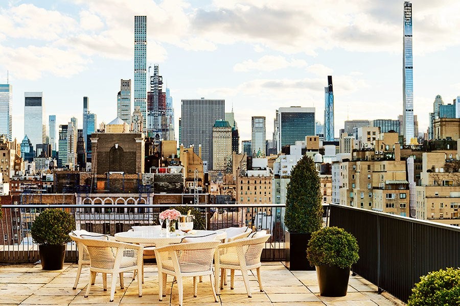 Rooftop patio with a round white table surrounded by six wicker chairs, decorated with a vase of pink flowers and a small plant. The patio has large potted shrubs and overlooks a city skyline with numerous tall buildings under a partly cloudy sky.