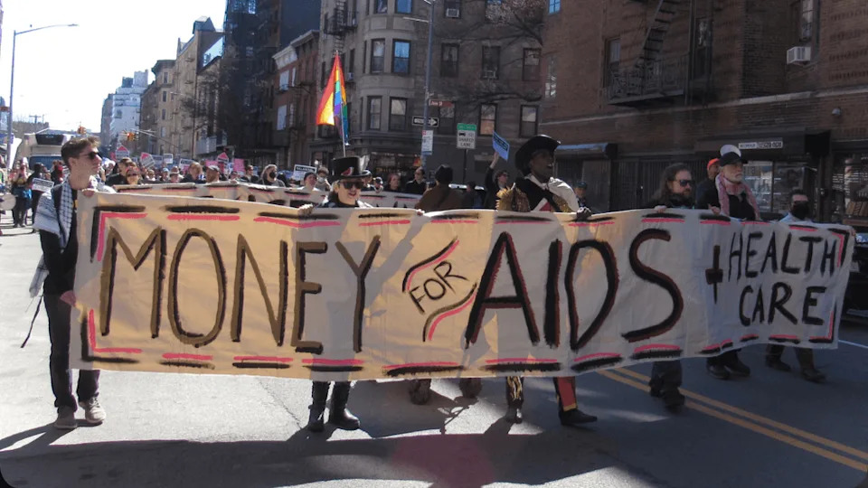 Protesters carry a banner through a street that reads "Money for AIDS and Health Care."