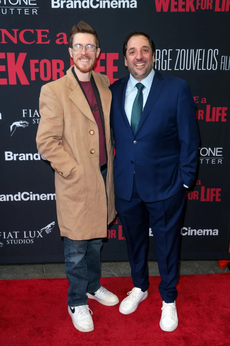 Two men in long coats and tennis shoes posing on red carpet. 