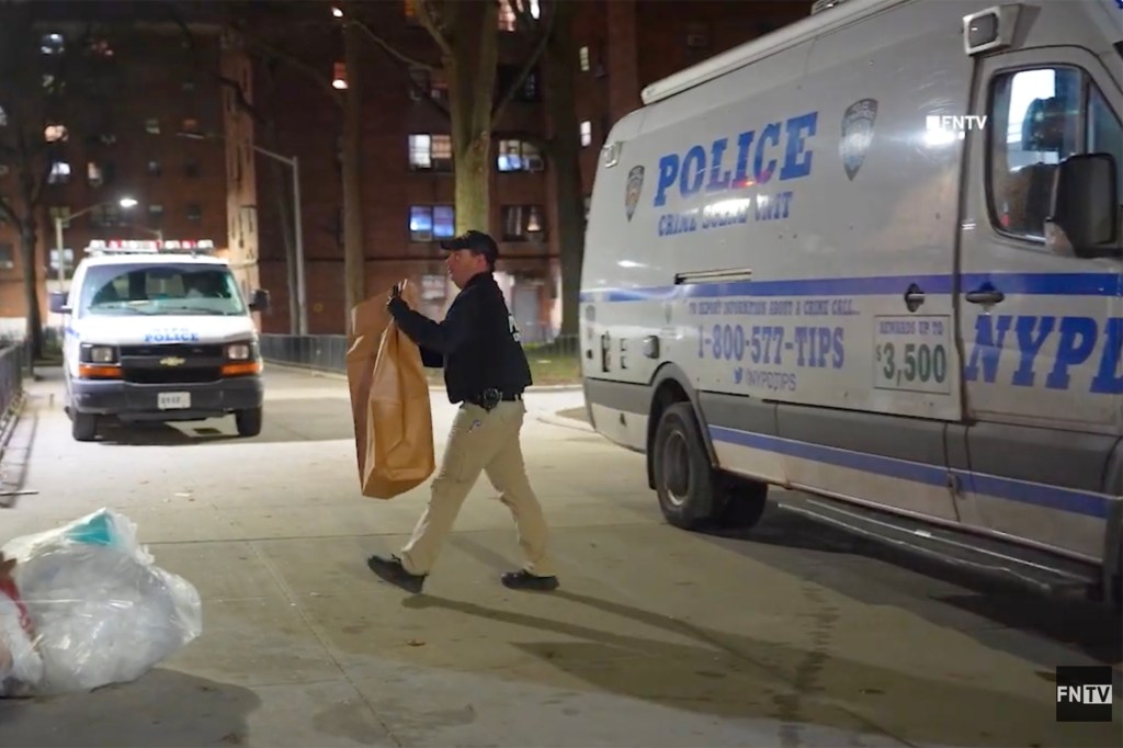 A police officer carrying a large brown bag walks past a police crime scene unit van at night.